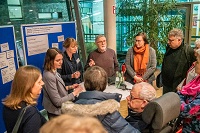 Menschen verschiedenen Alters stehen diskutierend um einen Stehtisch und vor Pinnwänden, auf denen ihre Beiträge von den Moderatorinnen fest gehalten werden; im Hintergrund sind Glaswände und Pflanzen zu sehen.