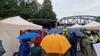 Das Foto zeigt im Hintergrund eine Bühne und im Vordergrund viele Menschen, die mit Regenjacken und Regenschirmen vor der Bühne stehen.