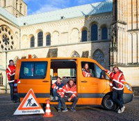 Fünf junge Leute in orangefarbigen Arbeitsanoraks an einem orangefarbigen Kleintransporter, davor ein Warnschild mit der Aufschrift  'Vermessung'.