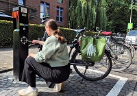 Auf dem Foto ist eine der drei neuen Radservicestationen abgebildet, hier an der Bismarckallee auf Höhe des Jugendgästehauses am Aasee. Die Radservicestation umfasst umfangreiches Werkzeug, um beispielsweise an einem Fahrrad Schrauben festziehen zu können und Luft aufzupumpen. Vor der Station kniet eine Frau mit ihrem Fahrrad, die gerade die Station in Anspruch nimmt, um Luft an ihrem Fahrrad aufzupumpen.
