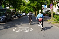 Abgebildet ist die Fahrradstraße Waldeyer- und Schmeddingstraße, auf der sich Radfahrende befinden, links und rechts am Fahrbandrand parken Autos, daneben kommt der Fußweg und angrenzende Wohnhäuser.