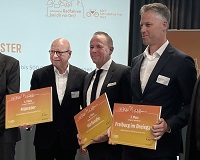 Bildmitte stehen 3 Männer im Anzug und weißem Hemd und halten die Preisverleihung in Form eines Schreibens - oranger Hintergrund - in der Hand.