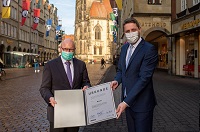 Zwei Männer mit Mund-Nasen-Schutz, bekleidet mit Anzug und Krawatte, schauen in die Kamera und halten die Urkunde in den Händen Richtung Kamera gerichtet. Im Hintergrund die Giebelhäuser in Münster