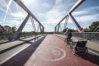 Brücke rot asphaltiert und das Piktogramm Fahrrad auf der Straße, auf der rechten Fahrradstraße fährt eine Frau ein Lastenrad.