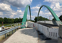 Im Hintergrund die Stahlbrücke mit grünen Bögen. Im Vordergrund Absperrbaken am Gehweg.