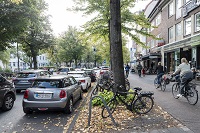Rechts im Bild Fahrradfahrende, links im bild parkende und fahrende Autos, mittig Baumbewuchs.