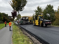 Asphaltiermaschinen stehen auf der neu asphaltierten Straße. Links auf dem angrenzenden Geh- und Radweg stehen mehrere Personen mit Warnkleidung und schauen auf die Straße.