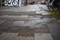 Auf dem Bild: Defekte Gehwegplatten und asphaltierte Flecken, rechter Bildrand geparkte Fahrräder dessen Hinterräder im Bild stehen. Oben links im Bild: weißes EBike steht im Schaufenster.