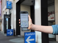 Vor einer Einfahrt zu einem Parkhaus ragt ein Arm von rechts ins mittig Bild, der ein Smartphone in der Hand hält, auf dem die neue Webseite zu sehen ist.