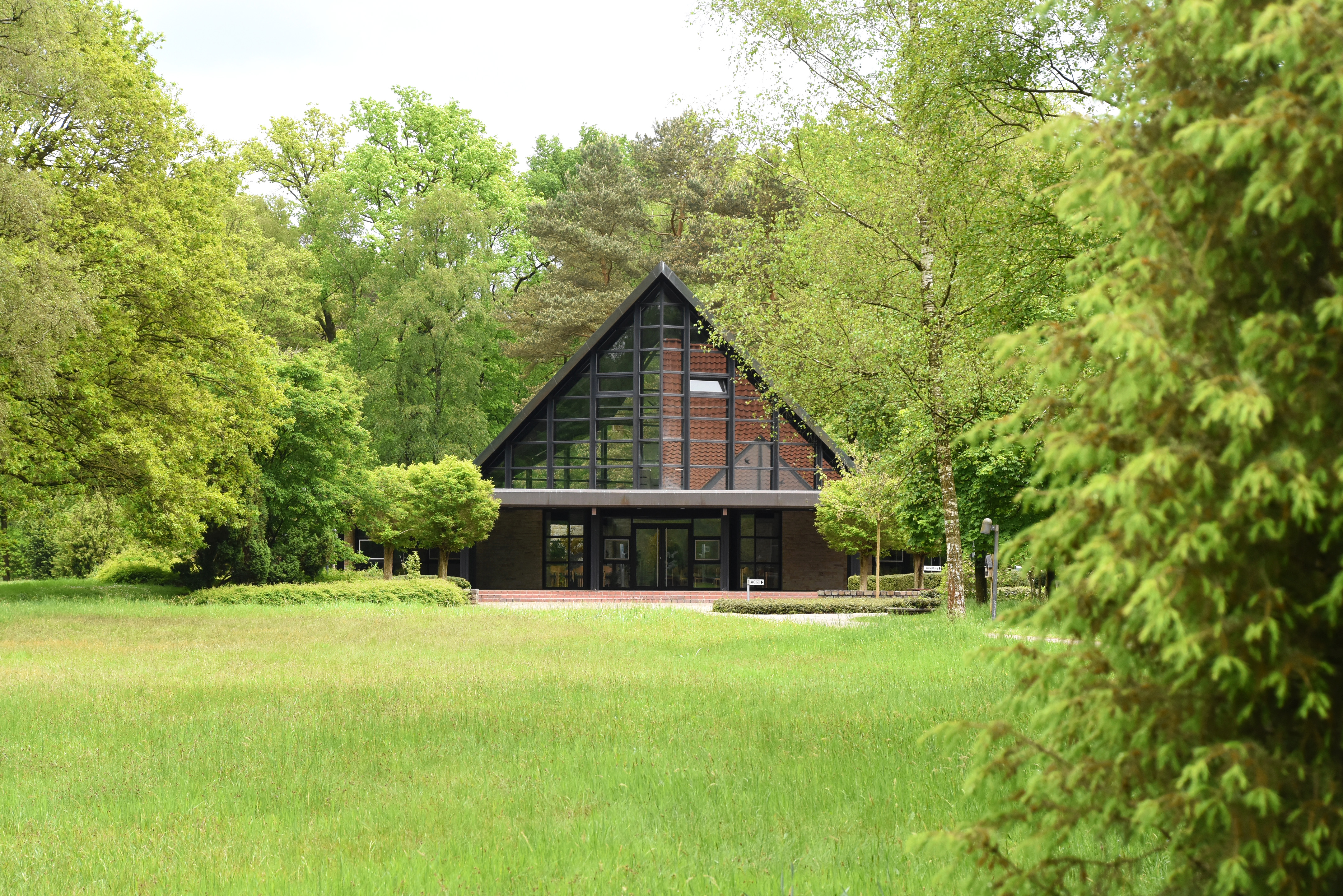 Im Hintergrund die Trauerhalle des Friedhofs Lauheide, im Vordergrund Büsche und eine Wiese erstreckt sich bis zur Trauerhalle.