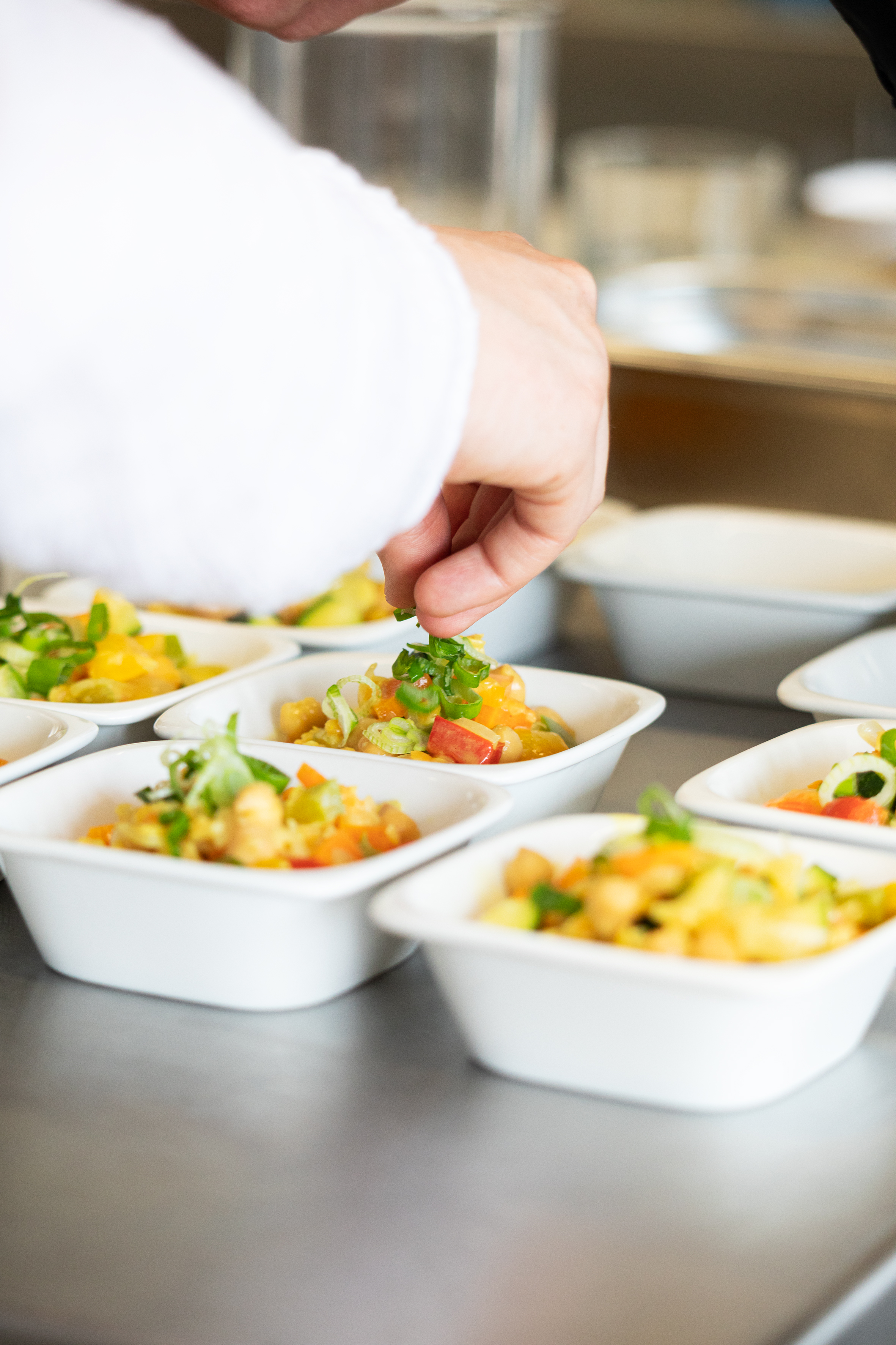 Mehrere Schälchen mit Essen auf einer Arbeitsplatte. Eine Hand verteilt darüber geschnittene Frühlingszwiebeln.