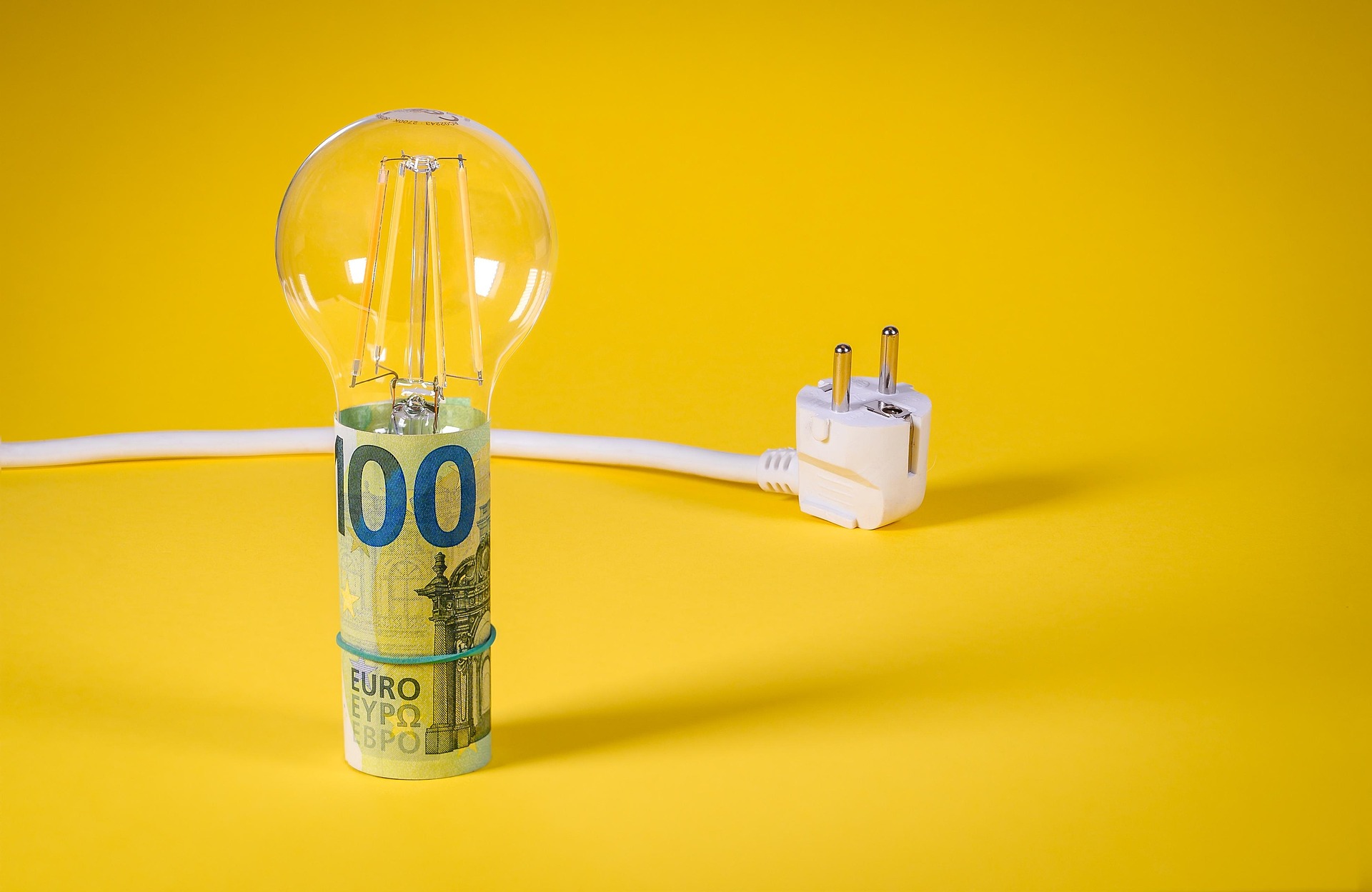 Auf knallgelben Grund liegt ein Stromkabel mit Stecker und in einer stehenden Geldrolle aus einem 100-Euro-Schein steckt eine Glühbirne