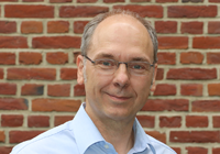 Ein Porträtfoto von Markus Wohlgemuth, Energieberater bei der Innovation City Management GmbH, vor einer Backsteinmauer. Er trägt ein blaues Hemd, Brille und hat eine Halbglatze.