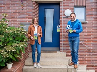 Ramona Leusch und Georg Reinhardt stehen lächelnd vor einer Haustür und halten Broschüren in die Kamera