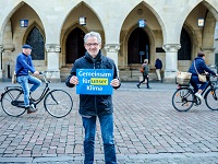 Klimadezernent Matthias Peck steht vor dem Rathaus und hält ein Plakat mit der Aufschrift "Gemeinsam für unser Klima". Im Hintergrund sind Radfahrende zu sehen.