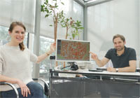 Eine Frau und ein Mann an einem Schreibtisch zeigen auf einen großen Computer-Bildschirm, der das Kataster anzeigt.