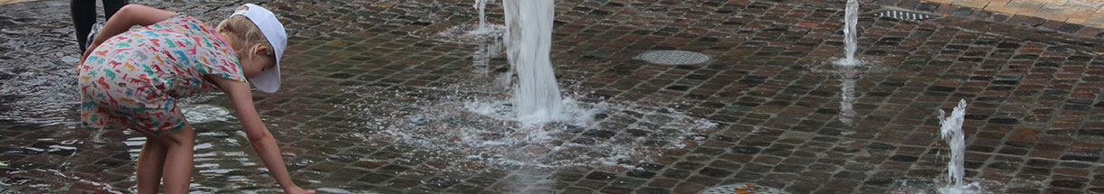 Ein Kind beugt sich über eine flache Wasserkuhle mit kleinen Springbrunnen.