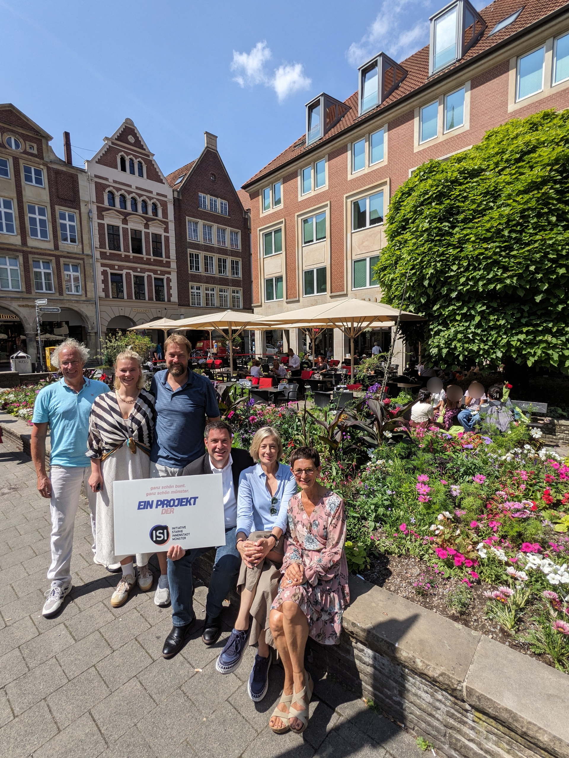 Zwei Frauen und ein Mann sitzen auf einer Mauer vor einem Blumenbeet. Der Mann hält ein Schild mit dem Logo der Initiative Starke Innenstadt. Dahinter stehen zwei Männer und eine Frau.