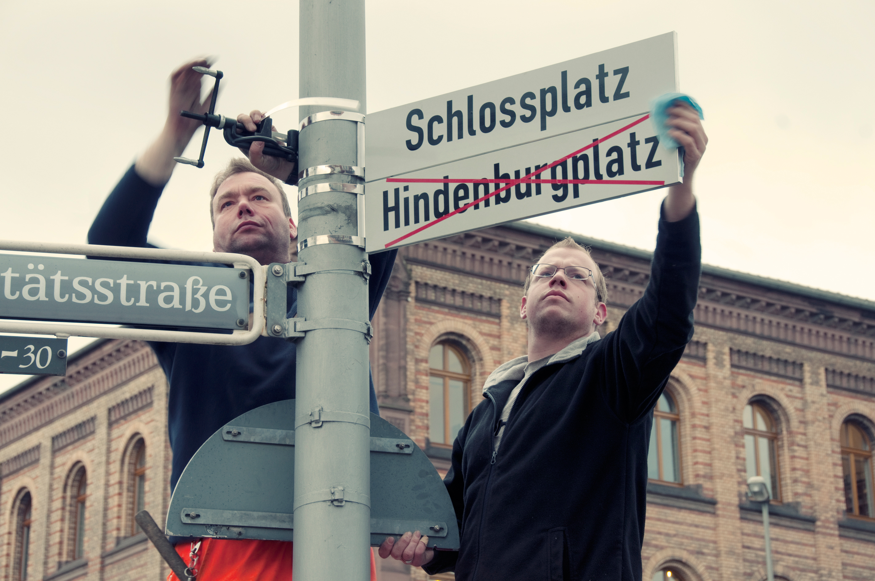 Zwei Männer tauschen Straßenschilder gegeneinander aus, Schlossplatz neu, Hindenburgplatz durchgestrichen