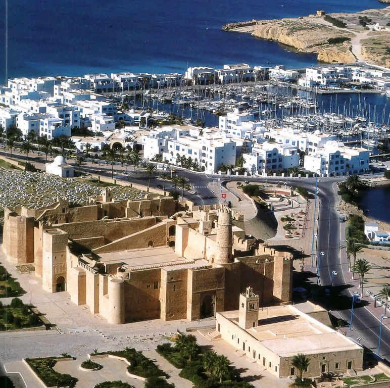 Blick auf die Stadt Monastir in Tunesien, es zeigt im Vordergrund eine historische Burganlage mit Palmengarten, in der Mitte weiße zwei bis drei geschossige Wohnhäuser, dahinter einen Jachthafen und ein Stück Mittelmeerküste