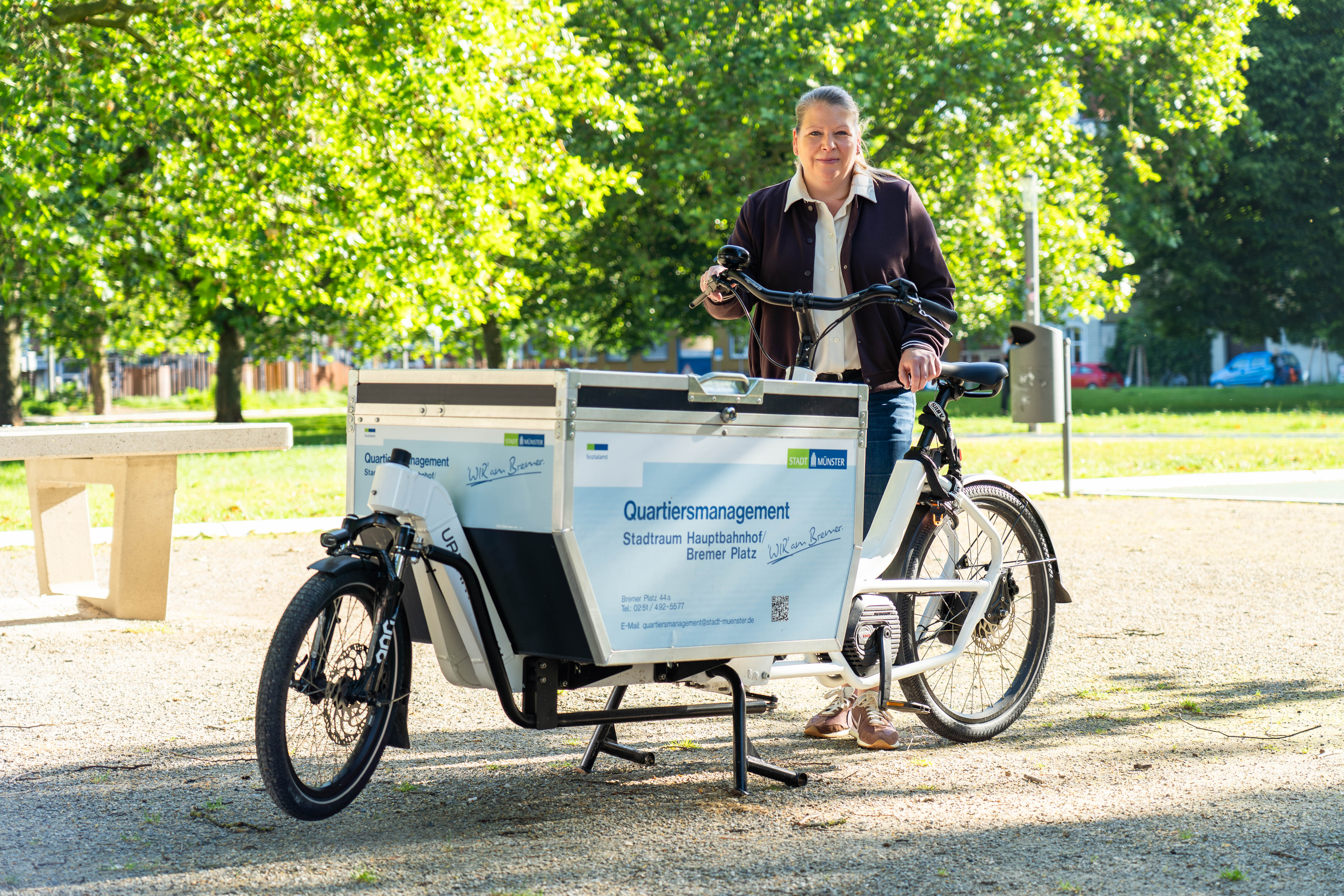 Quartiersmanagerin Judith Döpker mit dem Info-Lastenrad auf dem Bremer Platz, im Hintergrund die Grünfläche mit Spielgerät und Bäumen