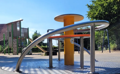 Spielplatz mit modernem, barrierefreiem Karussell aus Metall und Holz, im Hintergrund Klettergerüst und Schaukeln unter blauem Himmel