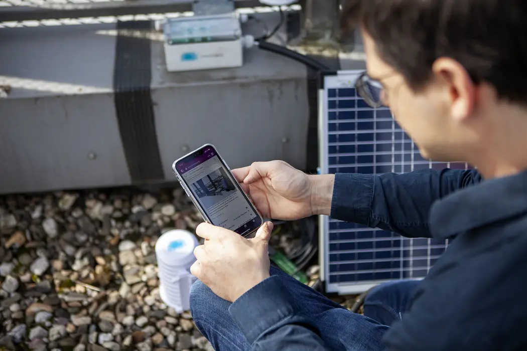 Eine männliche Person mittleren Alters schaut auf ein Smartphone, im Hintergrund sind Messgeräte und Solarzellen zu sehen, offenbar befindet sie sich auf dem Flachdach eines Gebäudessich auf 