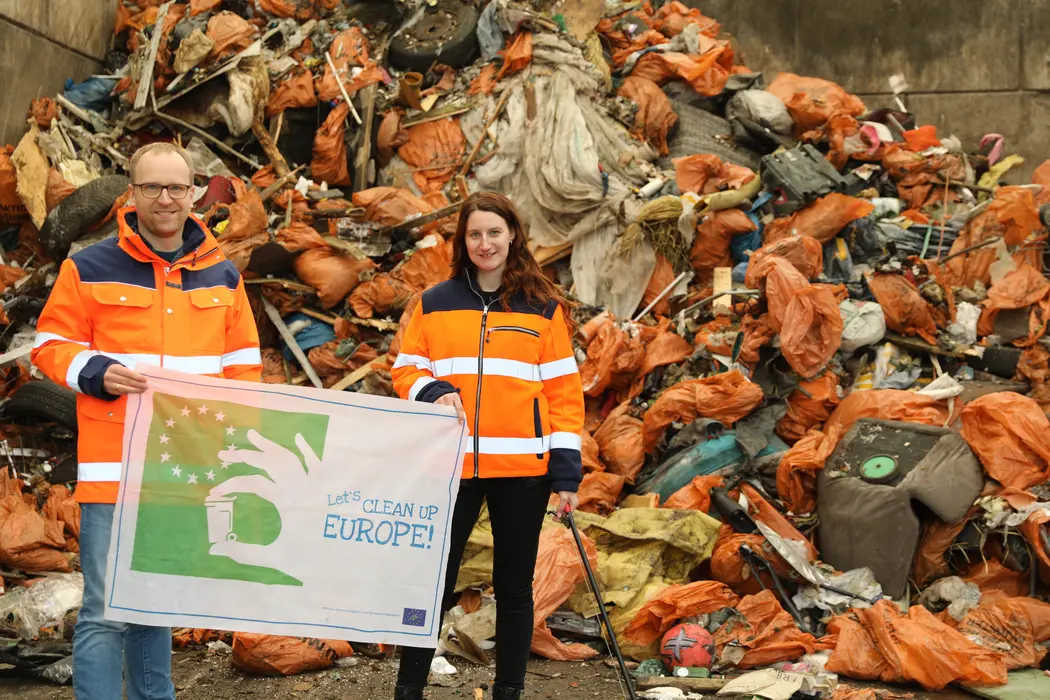 Betriebsleiter Dr. Baumkötter und Maren Wolff von den Abfallwirtschaftsbetrieben präsentieren die Bilanz der Aktion „Sauberes Münster 2026“, sie halten dabei ein Plakat mit der englischsprachigen Aufschrift "Wir machen Europa sauber", im Hintergrund ein großer Berg voller orangefarbener Sammelsäcke