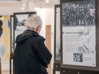 Zwei Menschen stehen vor Stahl-Paneelen, auf denen die Ausstellungsplakate von "Einige waren Nachbarn" angebracht sind. Das vordere Plakat zeigt eine Gruppe an KZ-Aufseher*innen und hat den Titel "Das Unbekannte und das Unvorstellbare".