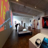 Ein heller Ausstellungsraum mit verschiedenen Exponaten, einer Projektion auf eine weiße Wand und einem Menschen, der sich vornüber beugt, um einen Ausstellungstext zu lesen.