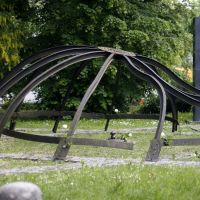 Ein Denkmal aus Metall, das an eine verbogene Kuppel erinnert, steht auf einem Davidstern aus Pflastersteinen auf einer Wiese. Dahinter sind eine Plakette und Bäume zu sehen.