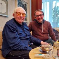 Zwei Männer sitzen auf einem Sofa, links der 97-jährige Margers Vestermanis, rechts Stefan Querl von der Villa ten Hompel. Beide blicken in die Kamera.