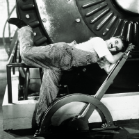 Charlie Chaplin hält einen großen Hebel mit beiden Händen fest gepackt, ein Bein hat er hinter sich in die Höhe gestreckt, als würde er von dem Schwung mitgerissen werden. Im Hintergrund ist Industriemaschinerie zu sehen.