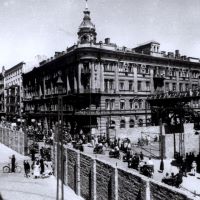 In einem historischen Schwarz-weiß-Foto trennt eine Mauer eine Straße mit wenigen Menschen von einem Stadtteil, in dem viele Menschen zu Fuß, mit Fahrrädern oder in Fuhrwerken unterwegs sind. Fünfstöckige Häuser säumen diese Straße im Warschauer Ghetto.