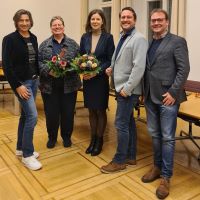 Ein Gruppenbild mit fünf Personen, die in die Kamera lächeln. Zwei der Frauen haben Blumensträuße in der Hand. Sie befinden sich in einem Raum mit Parkett