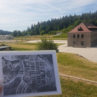 Im Vordergrund hält die Fotografin eine schwarzweiße Luftbildaufnahme hoch, auf der viele Strukturen von Häftlingsbarracken und anderen Gebäuden des Konzentrationslagers zu sehen sind. Im Hintergrund sieht man einen Teil des Geländes der Gedenkstätte: zwei größere Gebäude, einen Platz und Wald.