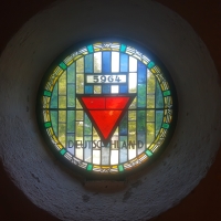 Ein rundes Glasmosaikfenster, in dessen Mitte ein roter Winkel steht. Unter ihm steht der Schriftzug ‚Deutschland‘, darüber die Zahl 5964 für die in dem KZ verstorbenen Deutschen.