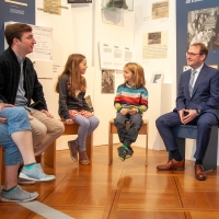 Mehrere Kinder und Erwachsene sitzen nebeneinander auf Hockern in der Dauerausstellung der Villa ten Hompel und unterhalten sich.