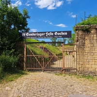 Über einem verwitterten Metalltor steht ein schwarzes Schild, auf dem geschrieben steht: 'SS Sonderlager Feste Goeben'