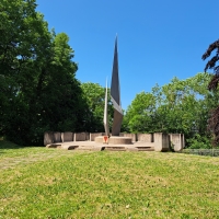 Auf einem Rasen umringt mit Bäumen steht ein Denkmal auf einer Fläche aus Stein. Es besteht aus zwei Spitzen, die in den Himmel ragen.