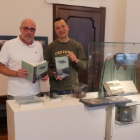 Thomas Köhler und Peter Römer stehen nebeneinander neben Vitrinen in der Ausstellung, halten je ein Exemplar des Sammelbandes hoch und lächeln in die Kamera.