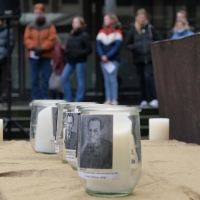 In einem Innenhof stehen mehrere Schüler*innen, einige haben die Hände vor sich zusammengefaltet. Im Vordergrund stehen mehrere Kerzen, auf denen die Namen und Bilder von Verfolgten des Holocaust aufgebracht sind.