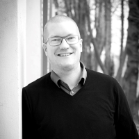 Ein Mann in Hemd, Pullover und Brille lehnt gegen einer Wand und lächelt in die Kamera.