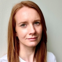 Eine Frau mit rotem Haar und weißem Shirt blickt mit einem Schmunzeln in die Kamera.