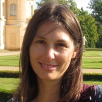 Eine Frau mit braunem Haar steht in einer Gartenanlage, ein helles Schloss im Hintergrund, und lächelt in die Kamera.