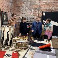 Zwei Männer und zwei Frauen stehen hinter einem Tisch, auf dem Sammlungsobjekte ausgebreitet sind. Dort liegen Trommeln, Kleidungsstücke und Abzeichen. An der Wand im Hintergrund hängt ein Bild Adolf Hitlers und eine SS-Fahne