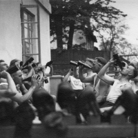 Auf einem historischen Foto sitzen Männer sich auf einer Veranda am Haus gegenüber und trinken aus Bierflaschen