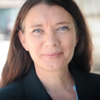 Eine Frau mit dunklen Haaren und Blazer schaut in die Kamera