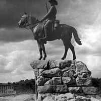 Unter einem bewölkten Himmel, der den größten Teil des Fotos einnimmt, steht eine Reiterstatue auf einem Sockel aus verschiedenen Steinen. Die Reiterfigur trägt ein Gewehr in einem Arm. Im Hintergrund ist ein zweistöckiges Gebäude zu erkennen.