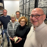 Fünf Menschen stehen zwischen zwei Archivregalen, die mit Archivkisten gefüllt sind, und blicken in die Kamera.